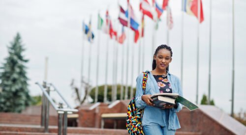 African student female posed with backpack and school items on yard of university, against flags of different countries.